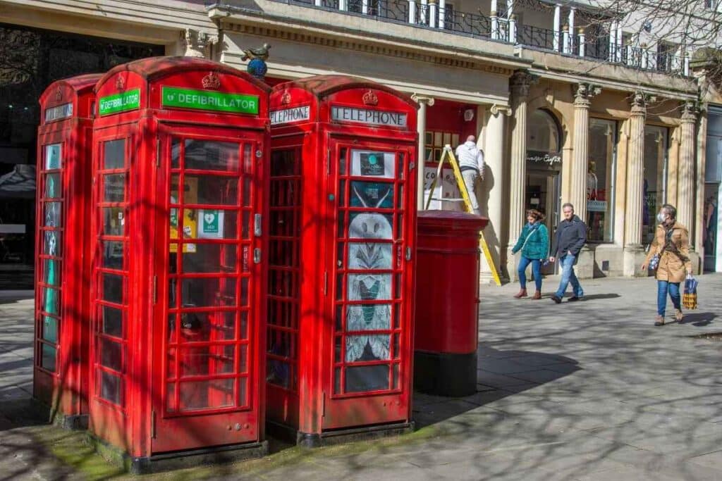 Three red telephone boxes on a high street, one with a defibrillator cabinet inside.