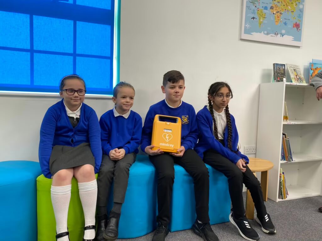 Group of school children holding an AED defibrillator, emphasizing the importance of having defibrillators in schools for emergency preparedness.