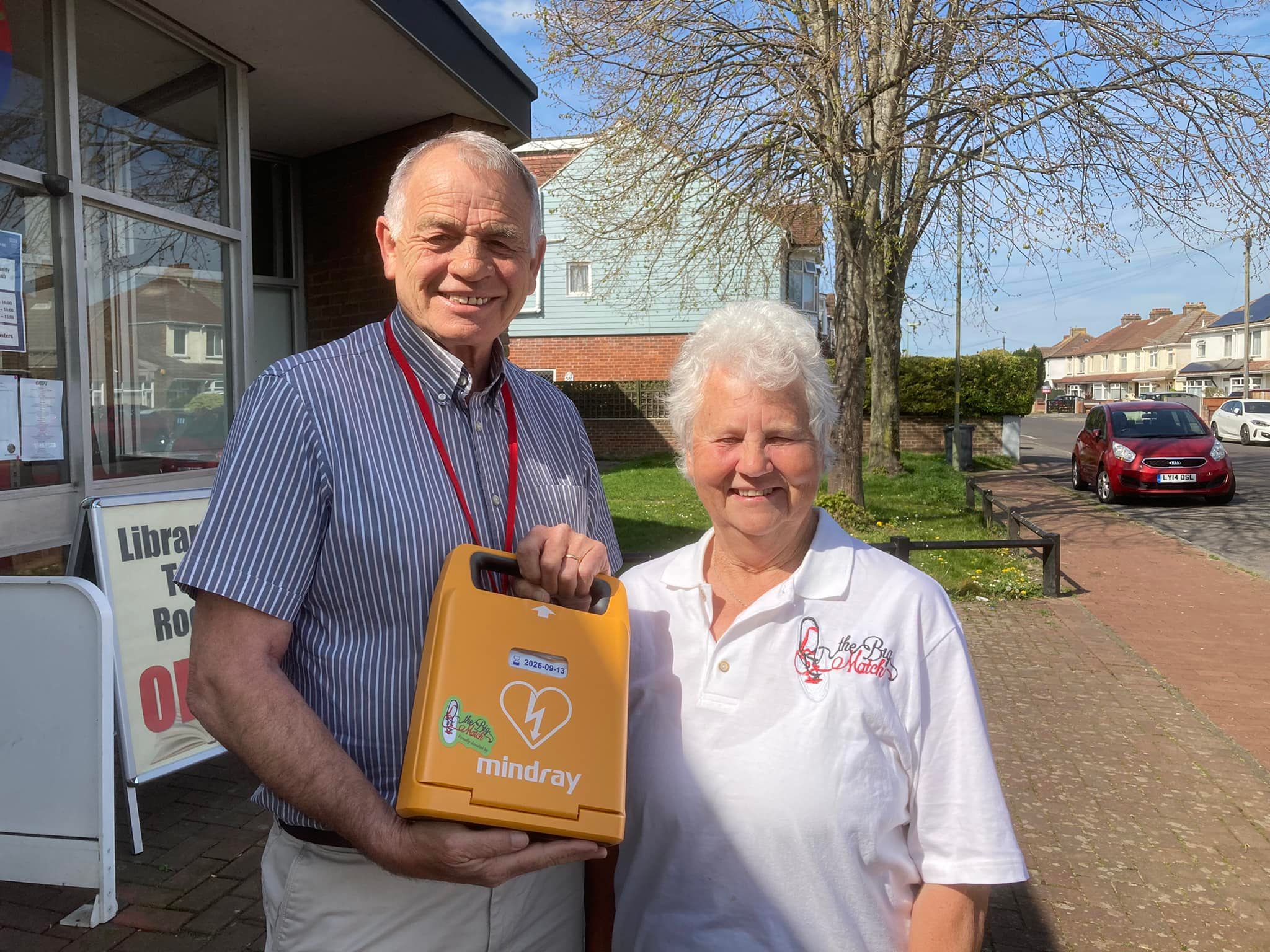 Two community members smile as they pose outside a library, holding a newly donated Mindray AED device for public use.