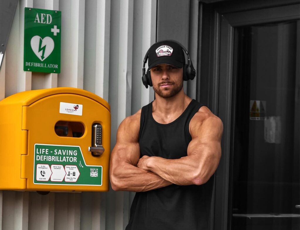 A fit man wearing a black tank top and headphones stands next to a life-saving defibrillator mounted outside a gym, emphasizing AED availability for emergencies.