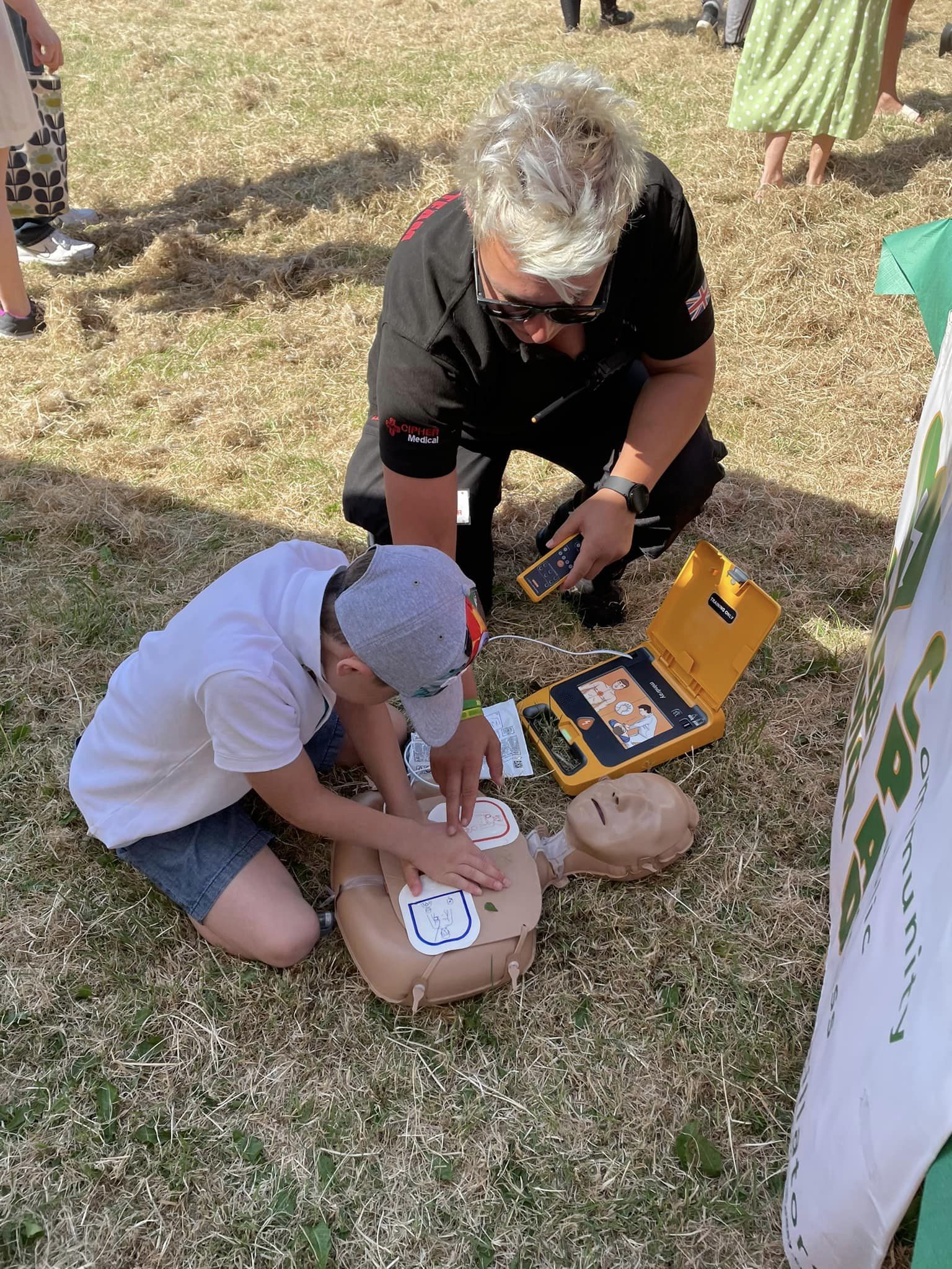 A child practicing CPR on a mannequin with the guidance of an instructor, using a defibrillator, during a community event.