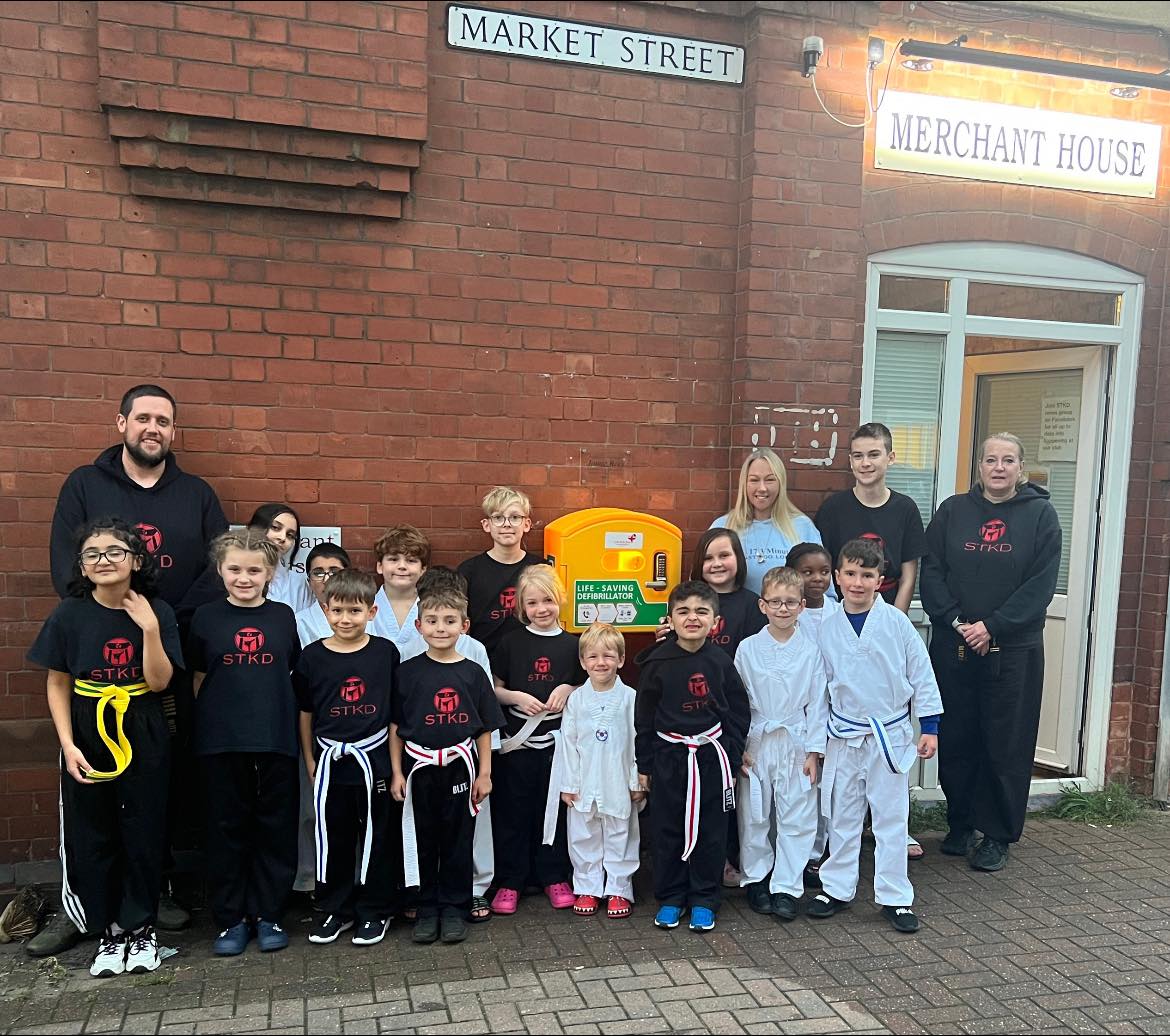 A group of children and adults from STKD martial arts standing in front of a defibrillator outside Merchant House on Market Street.
