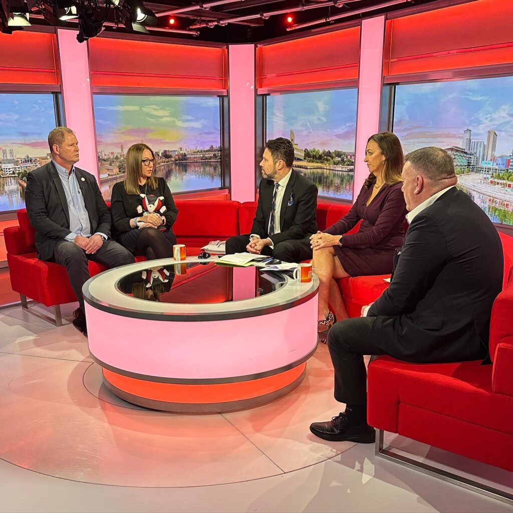 A group of four people sits on a red couch in a studio setting, engaged in a discussion with a host. The focus is on raising awareness about defibrillators in public spaces.
