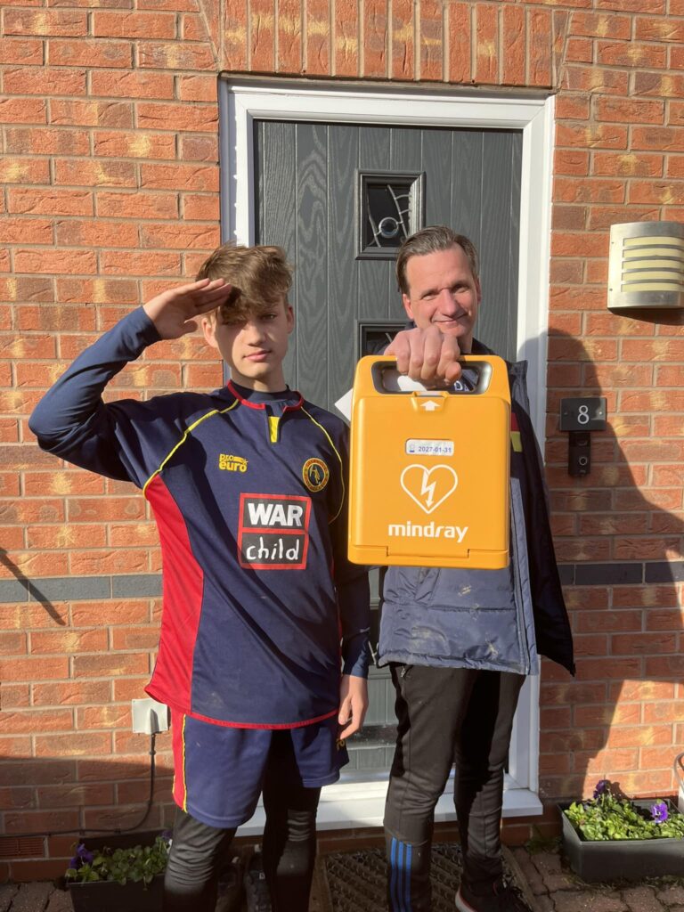 Two individuals standing outside a house, holding a Mindray defibrillator. The man is presenting the device while the boy salutes, emphasizing the importance of defibrillator access and awareness in public spaces for heart emergencies.