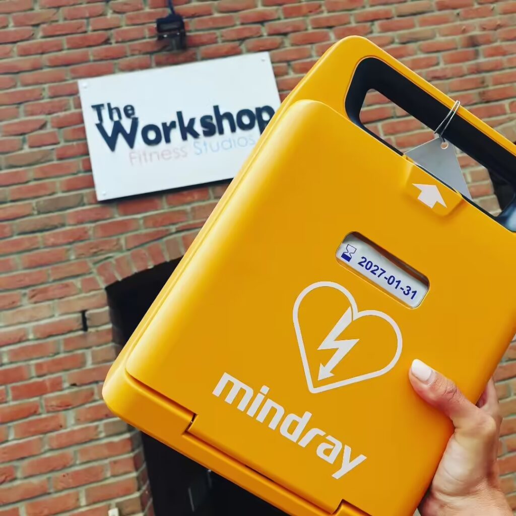 Mindray AED being held outside a fitness studio, with an expiration date of 2027 visible on the device.