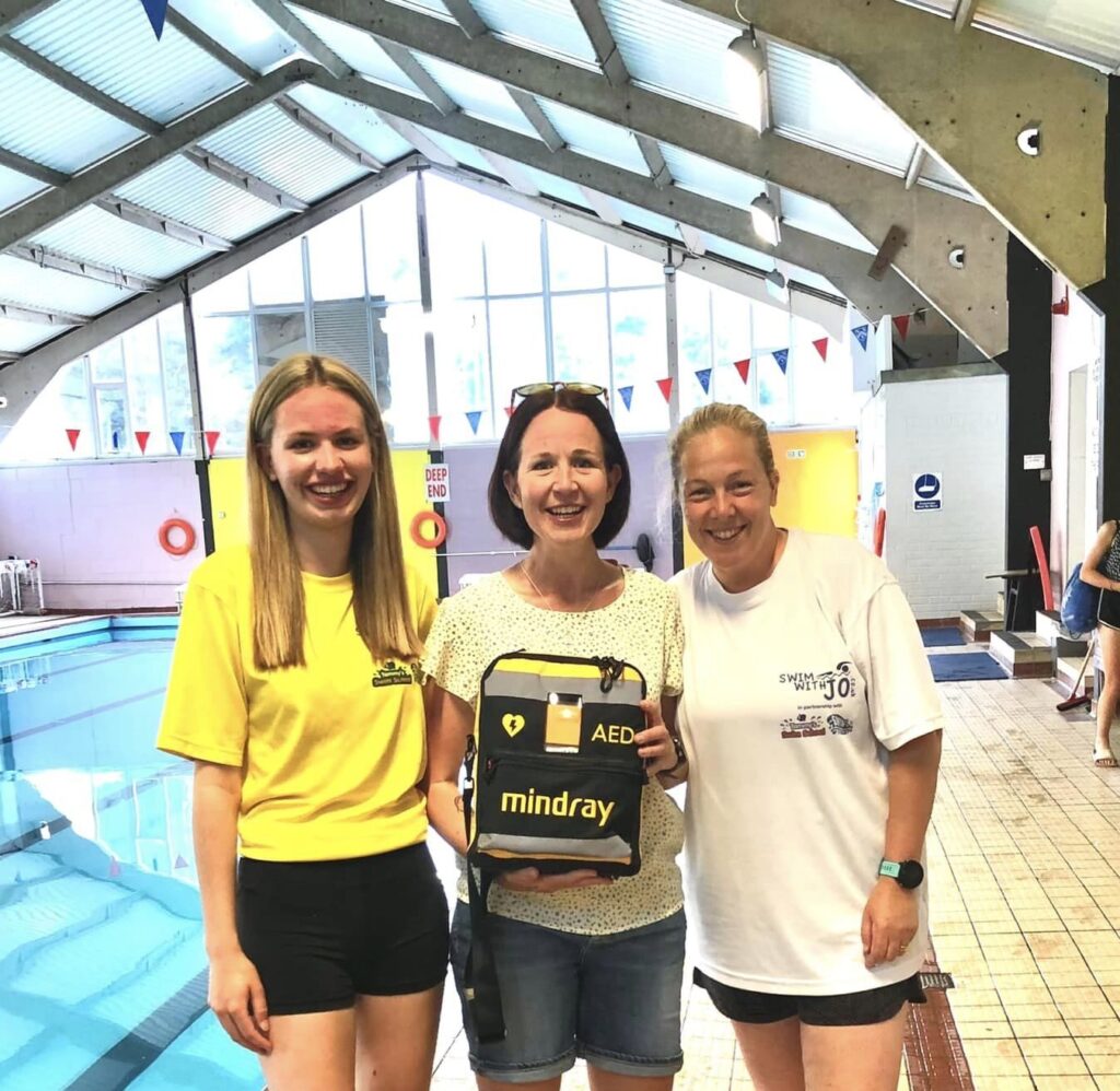 Three women proudly holding a Mindray AED defibrillator donated to a swimming pool for life-saving emergencies.