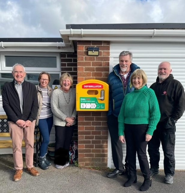 Community members standing in front of a defibrillator mounted on a wall, highlighting the importance of public access to AEDs in the UK, supported by Defib World