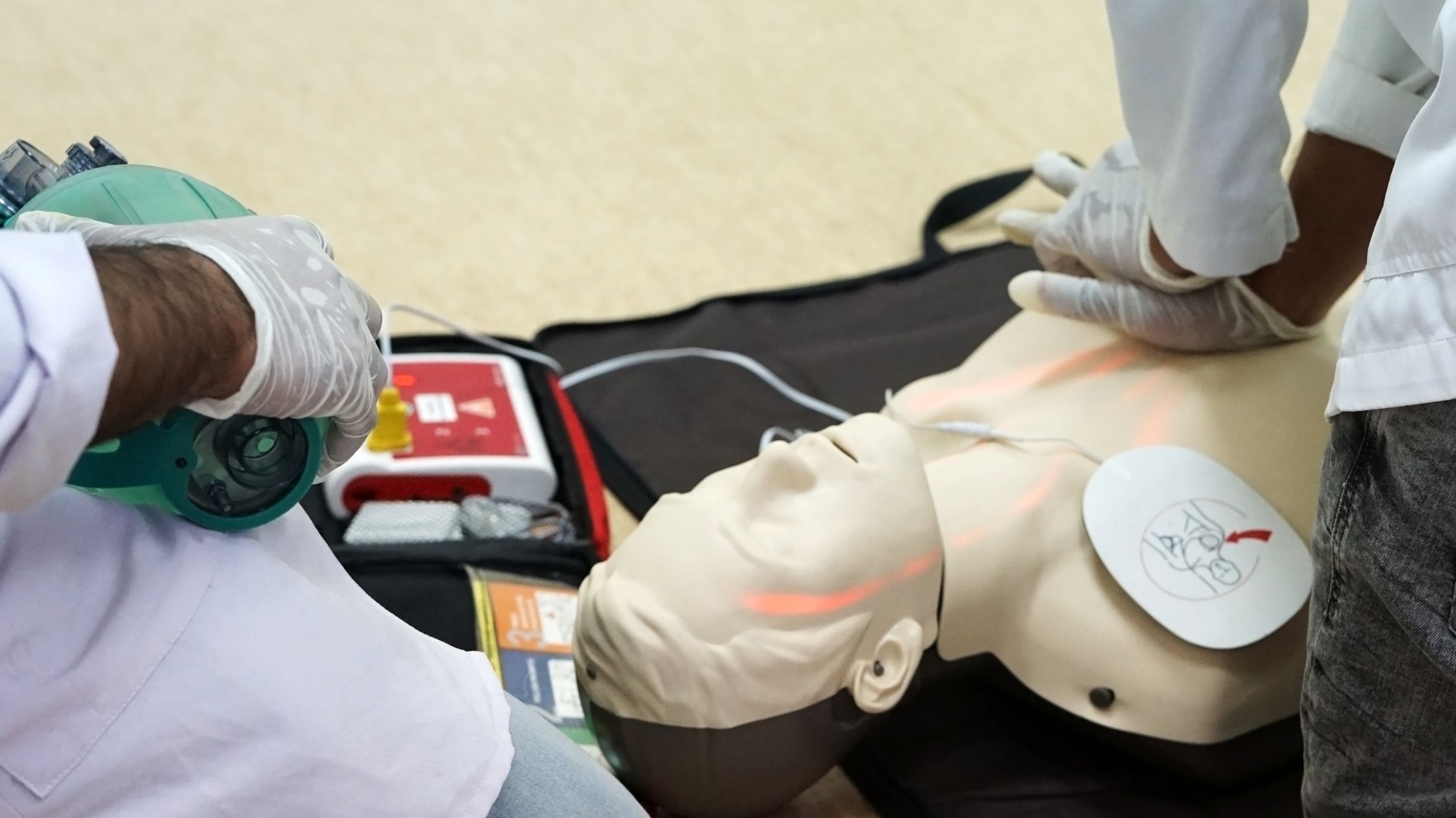A person performing CPR on a manikin with an AED device visible in the background.