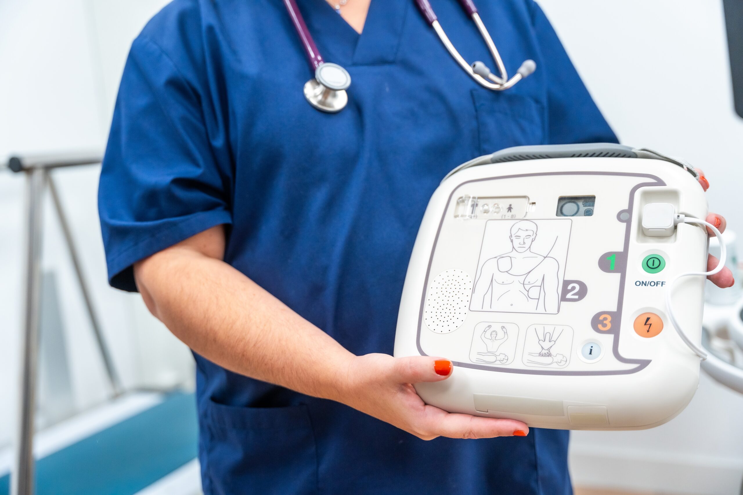 Medical professional holding an AED device, ready to be used in an emergency. The image highlights the design of the defibrillator, with clear instruction symbols for usage. It is used to emphasize the importance of having AED devices accessible in medical and emergency situations.