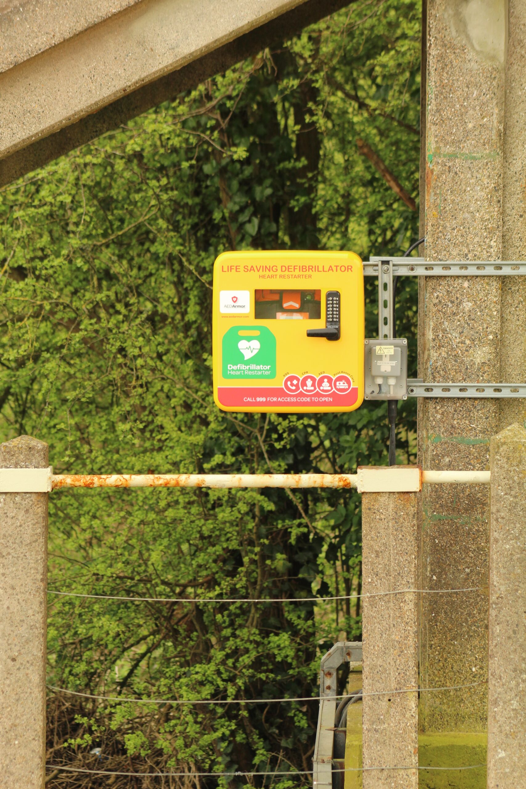 A bright yellow defibrillator mounted on a fence, showcasing AED placement in rural or less accessible areas, emphasizing its importance in all environments.